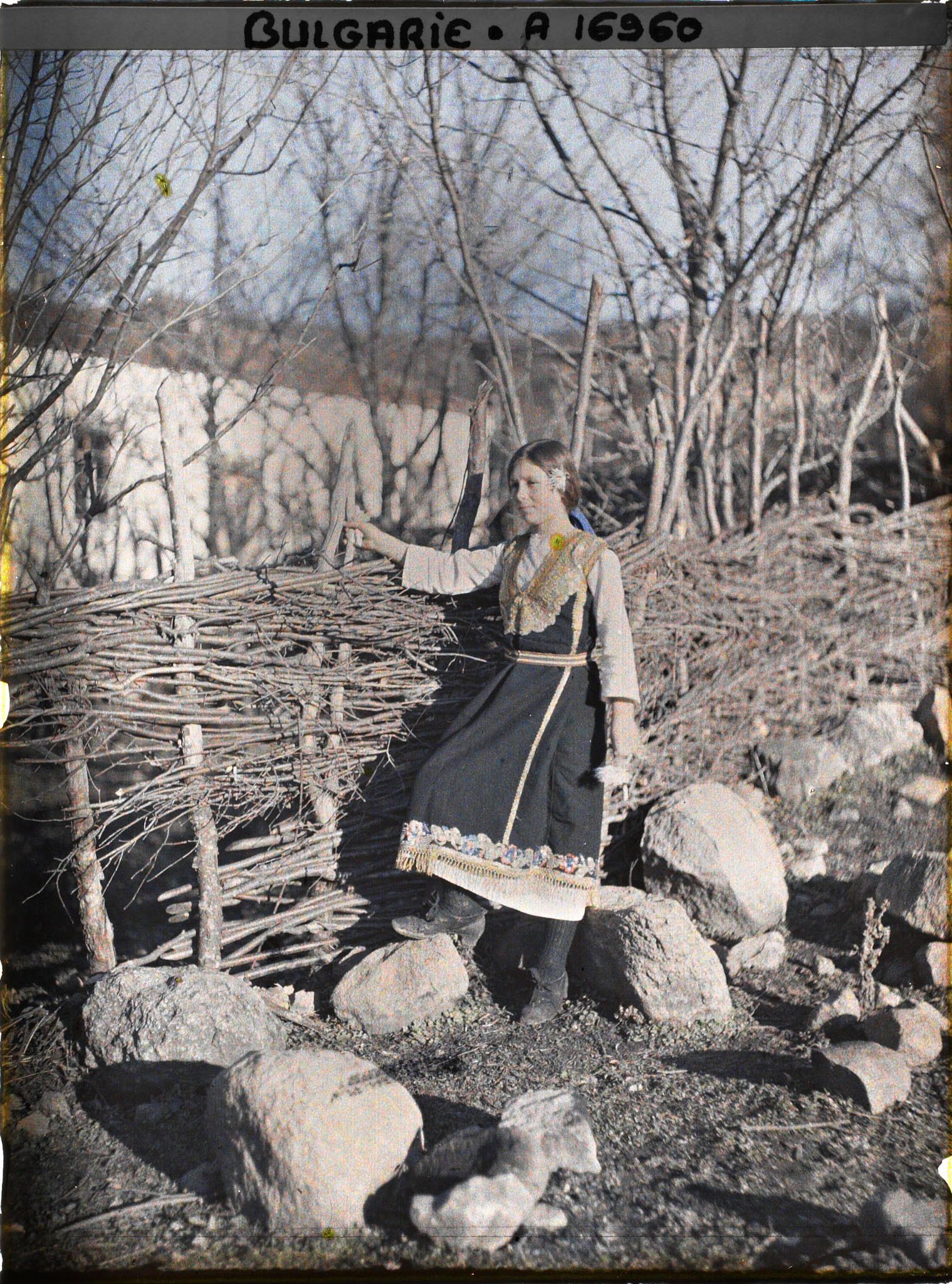 Image représentant Jeune fille, en robe brodée, devant un mur de petit bois avec à l'arrière-plan l'église