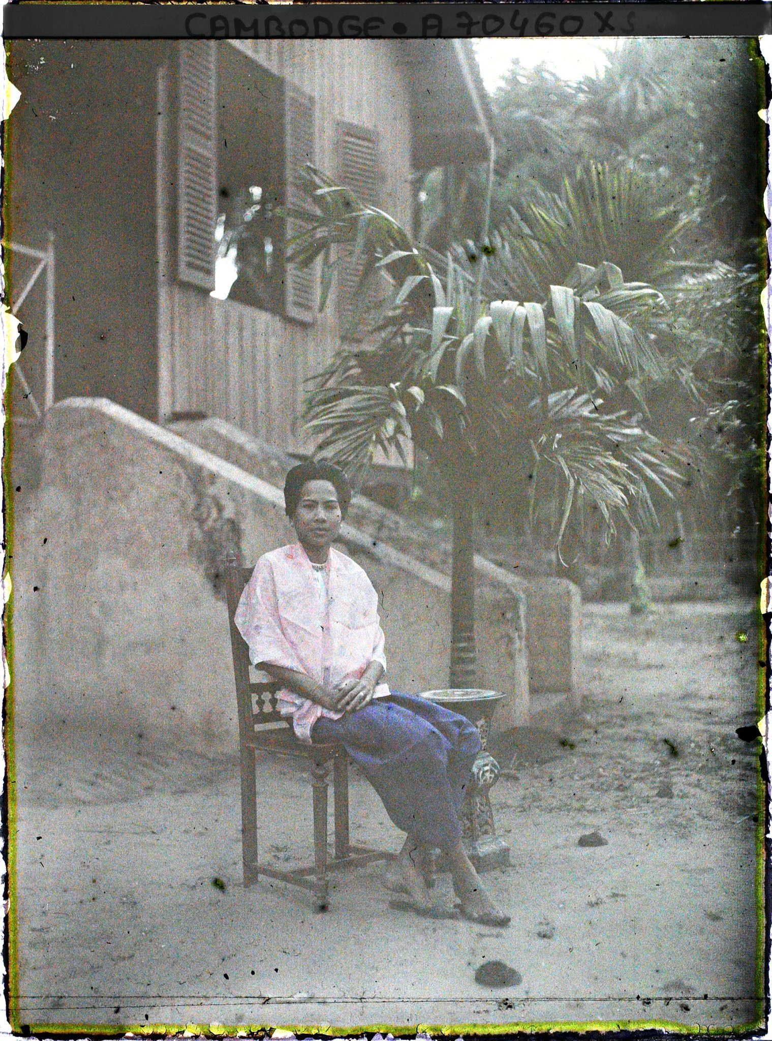 Image représentant Femme en sampot violet, assise sur une chaise de bois tournée devant le perron d'une maison