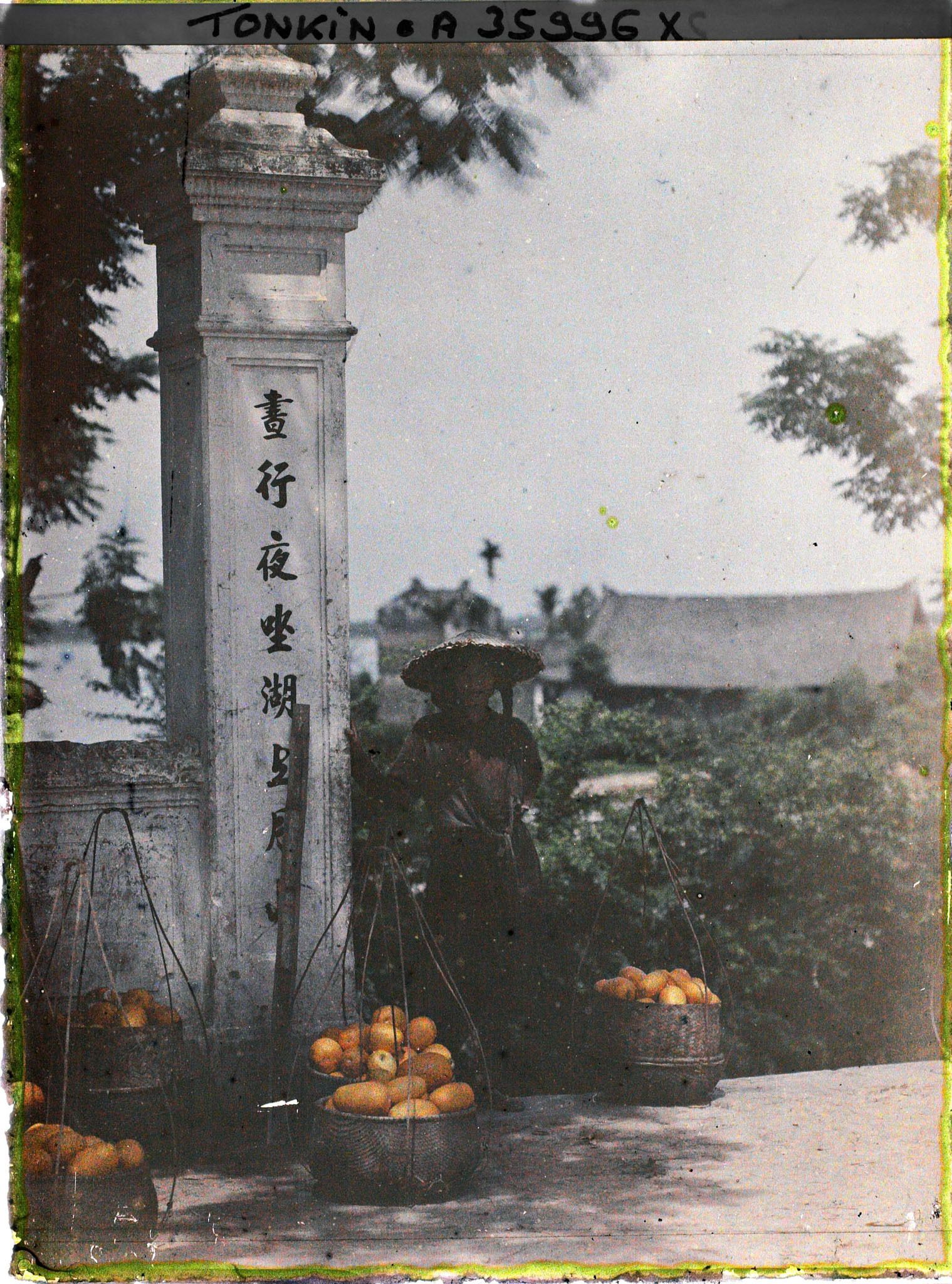 Image représentant Une femme annamite entourée de paniers de fruits (ou légumes), pose pour le photographe près d'un portique au temple Ngoc-so'n
