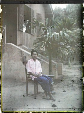 Image représentant Femme en sampot violet, assise sur une chaise de bois tournée devant le perron d'une maison
