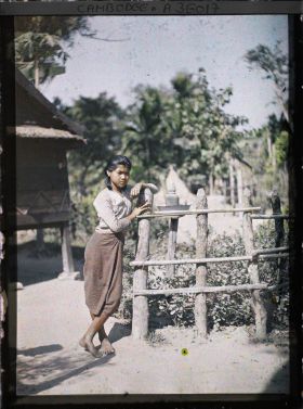 Image représentant Une jeune fille accoudée à une barrière, derrière laquelle on aperçoit une " maison des Esprits " (autel domestique)