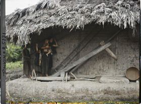 Image représentant Une femme accompagnée d'enfants actionnant au pied le pilon à blanchir le riz, dans une cour de ferme après la moisson du riz