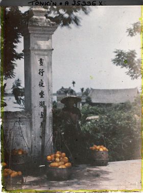 Image représentant Une femme annamite entourée de paniers de fruits (ou légumes), pose pour le photographe près d'un portique au temple Ngoc-so'n